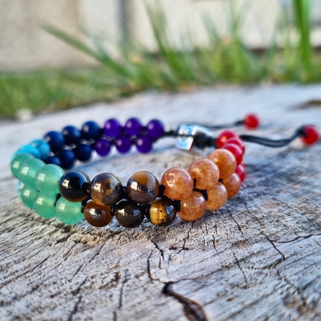 Handmade bracelet with 7 chakra crystal gemstones, featuring amethyst, lapis lazuli, blue aquamarine, green aventurine, tiger’s eye, orange stone, and red agate. The bracelet is on a piece of wood in nature.