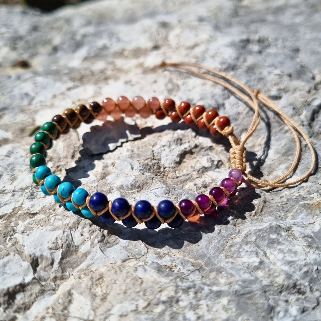 Handmade bracelet with 7 chakra crystal gemstones, featuring amethyst, lapis lazuli, turquoise,  malachite, tiger’s eye, sunstone, and red agate. The bracelet is positioned on a stone surface. 
