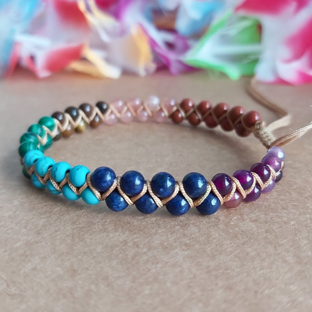 Handmade bracelet with 7 chakra crystal gemstones, featuring amethyst, lapis lazuli, turquoise,  malachite, tiger’s eye, sunstone, and red agate. The bracelet is positioned on a nice surface with colourful flowers in the background.